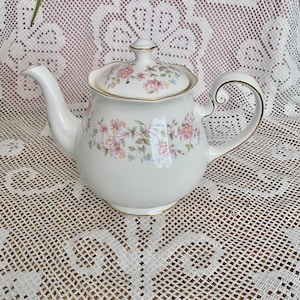 May include: A white porcelain teapot featuring a delicate floral pattern with pink and yellow flowers and green leaves. The teapot has a matching lid with a small knob, a curved handle, a tapered spout, and gold trim along the edges.