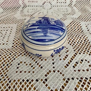 May include: A small, round ceramic trinket box with a lid. The lid features a blue and white illustration of a windmill and landscape. The box has a cream-coloured base with blue floral accents. The box is approximately 7.6 cm in diameter.