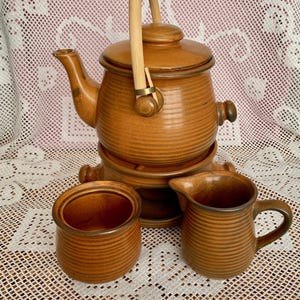 May include: A brown ceramic tea set with a teapot, creamer, and sugar bowl. The teapot has a wooden handle and lid. The set is on a white lace tablecloth. The teapot is approximately 25 cm tall.