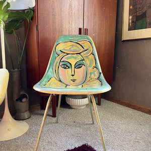 May include: A vintage green and yellow chair with a painted portrait of a woman's face. The chair has a metal frame and four legs.