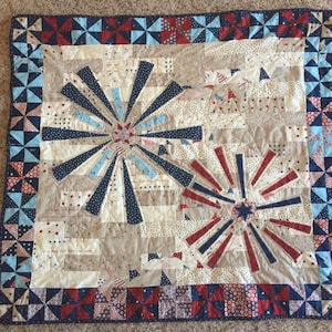 May include: A handmade quilt with a patriotic theme. The quilt features a central design of starbursts in red, white, and blue, surrounded by a border of pinwheel patterns. The quilt is approximately 48 inches by 48 inches.