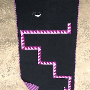 May include: A black quilt with a pink and white geometric design. The quilt has a white smiling face in the center.