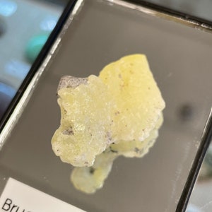 May include: A small, pale yellow brucite mineral specimen with a rough, crystalline texture. The mineral is displayed in a glass case with a label that reads "Bruceite $10".