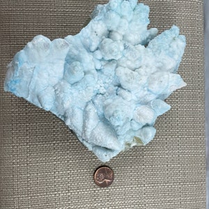 May include: A large, light blue, irregularly shaped rock with a bubbly, textured surface. A penny is placed next to the rock for scale.