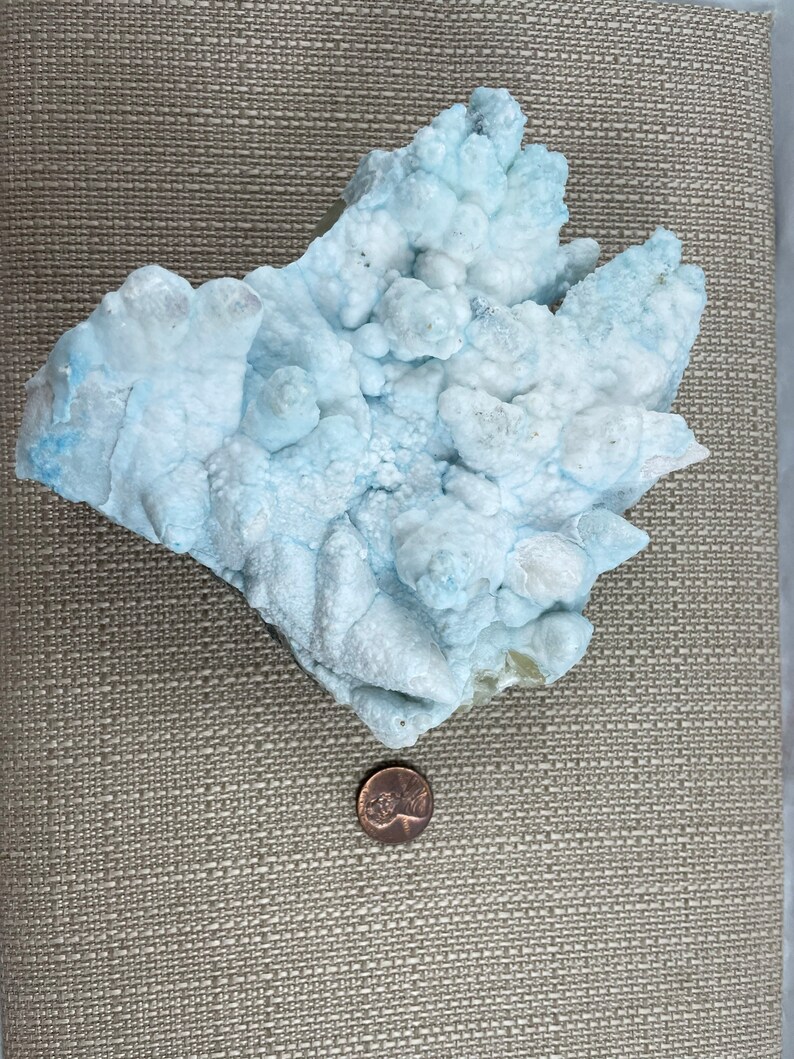 May include: A large, light blue, irregularly shaped rock with a bubbly, textured surface. A penny is placed next to the rock for scale.