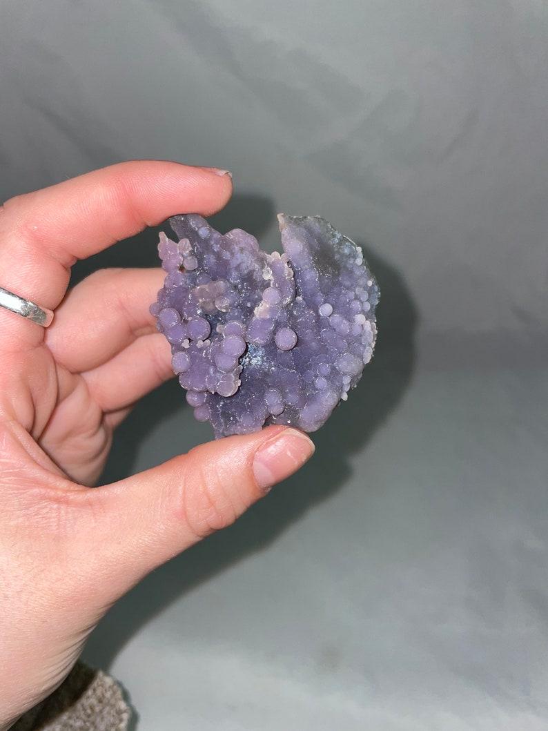 K&ouml;nnte beinhalten: Eine violette Achat-Geode mit einem Cluster aus kleinen, runden, violetten Achat-Kn&ouml;tchen. Die Geode ist eine nat&uuml;rliche Formation mit einer rauen, unregelm&auml;&szlig;igen Form.