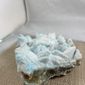 May include: A cluster of blue and white celestite crystals with a rough, natural texture. The crystals are arranged in a jagged, irregular shape, with some of the points broken off. The celestite is a light blue color, with some white areas. The crystals are set against a brown, textured background.
