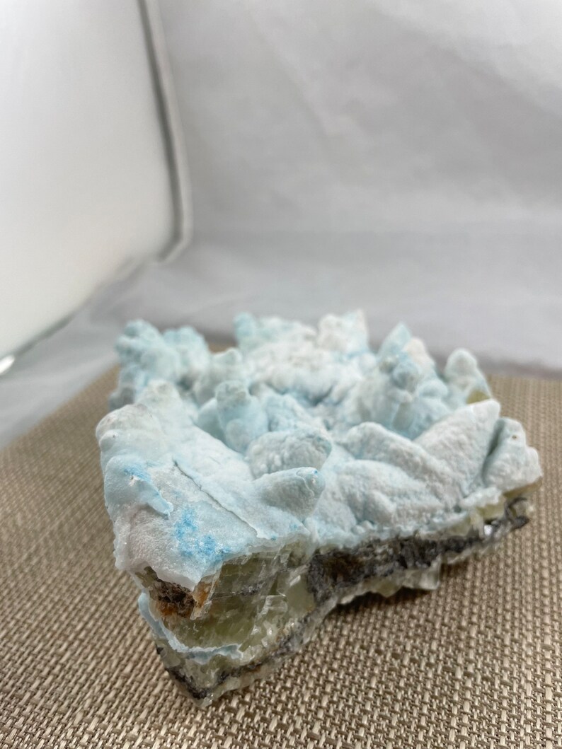 May include: A cluster of blue and white celestite crystals with a rough, natural texture. The crystals are arranged in a jagged, irregular shape, with some of the points broken off. The celestite is a light blue color, with some white areas. The crystals are set against a brown, textured background.
