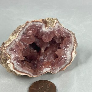 May include: A pink amethyst geode with a natural, rough exterior and a hollow interior lined with sparkling pink crystals. The geode is about the size of a US penny.