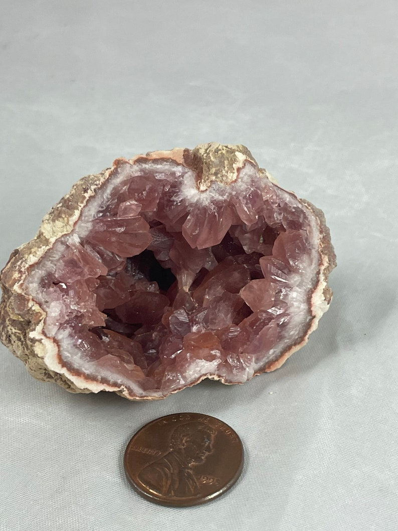 May include: A pink amethyst geode with a natural, rough exterior and a hollow interior lined with sparkling pink crystals. The geode is about the size of a US penny.
