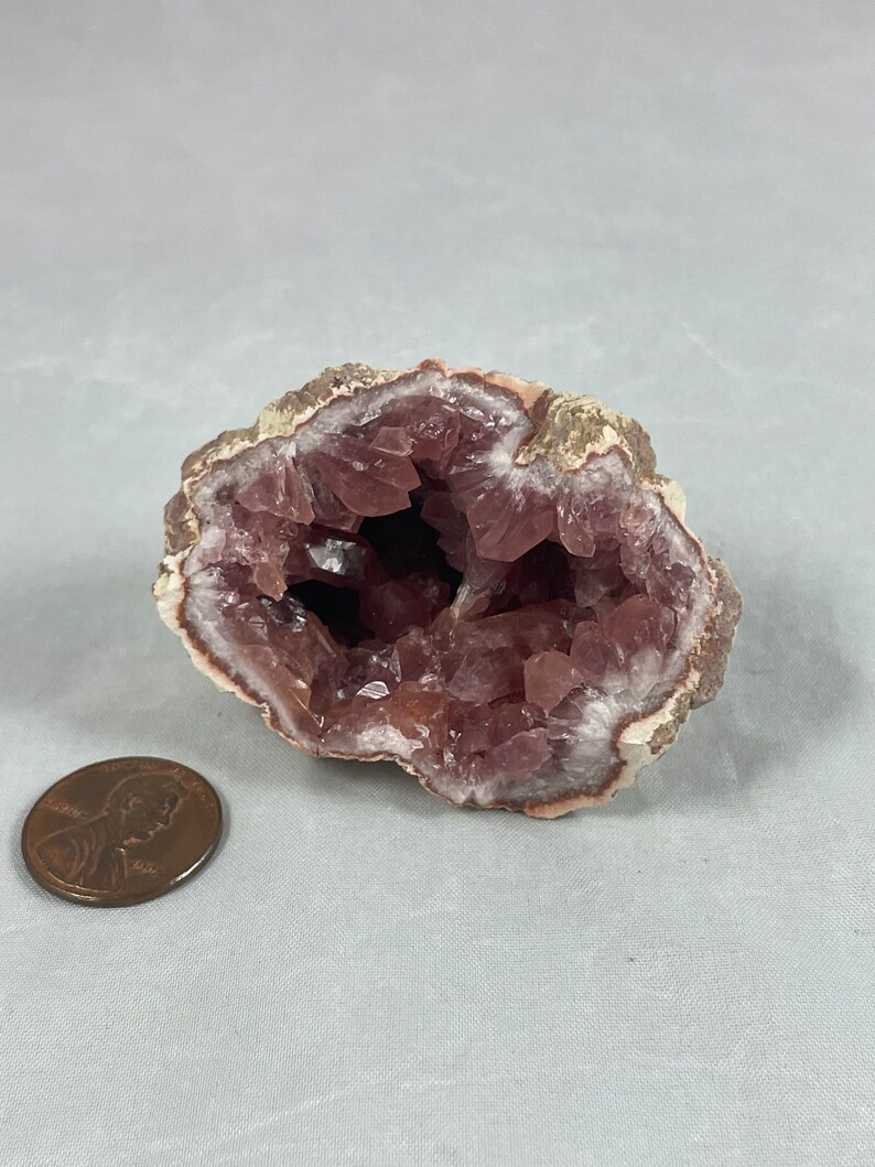 May include: A pink amethyst geode with a hollow center and a rough exterior. The geode is about the size of a US penny.