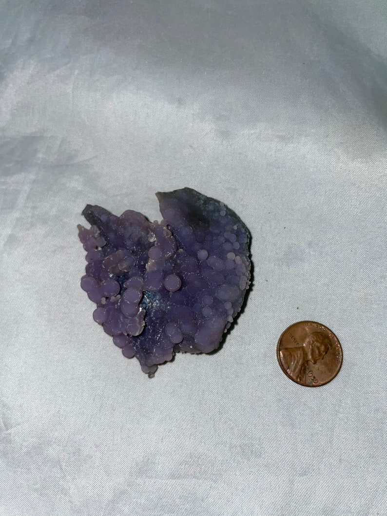 K&ouml;nnte beinhalten: Ein violetter und grauer Achat-Geode mit einer rauen, strukturierten Oberfl&auml;che. Der Geode ist mit kleinen, runden Beulen bedeckt. Ein Penny ist zur Gr&ouml;&szlig;enbestimmung enthalten.
