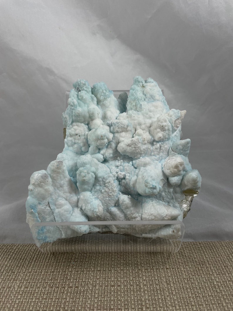 May include: A cluster of blue and white celestite crystals in a clear acrylic display case. The crystals have a rough, textured surface and are arranged in a random, organic pattern.