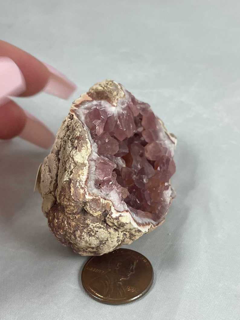 May include: A pink amethyst geode with a rough exterior and a hollow interior filled with sparkling pink crystals. The geode is about the size of a US penny.