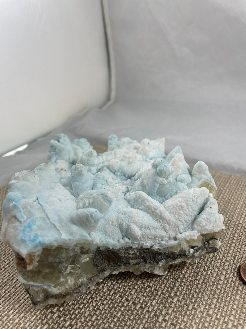 May include: A blue and white crystal cluster with a rough, textured surface. The crystal has a layered structure with a darker, gray layer at the bottom.
