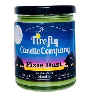 May include: A green glass jar candle with a black lid. The label says "Firefly Candle Company" and "Pixie Dust" with a moon and firefly graphic. The label also says "handmade in Hilton Head Island South Carolina".