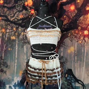 May include: A two-piece crochet set consisting of a white crop top and a matching skirt. The top features thin white straps and a ruffled beige trim. The skirt has a white base with layered brown and dark brown ruffled trim and a brown tie.