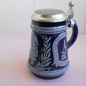 以下が含まれることがあります： ピューター製の蓋が付いた青と白のセラミック製のビアジョッキ。ジョッキには、ドイツの医師ロベルト・コッホの肖像画と、「ノーベル医学生理学賞」と「バクテリア」の文字が描かれています。