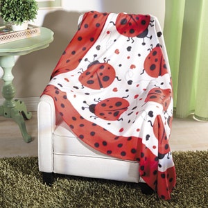 May include: A white blanket with a red border and ladybug pattern draped over a white armchair. The blanket features large red ladybugs with black spots and antennae, along with small black hearts.