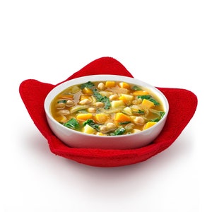 May include: A white bowl filled with vegetable soup, featuring chickpeas, squash, and greens. The bowl rests on a bright red, heart-shaped, fabric pot holder, set against a white background. The soup appears warm and inviting.