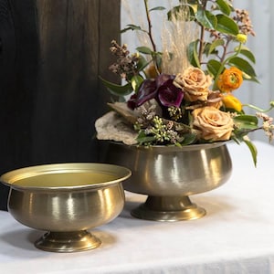 May include: Two antique brass bowls, one small and one large, sit on a white surface. The larger bowl holds a floral arrangement with peach roses, purple calla lilies, and greenery. The bowls have a textured finish and are on pedestal bases.