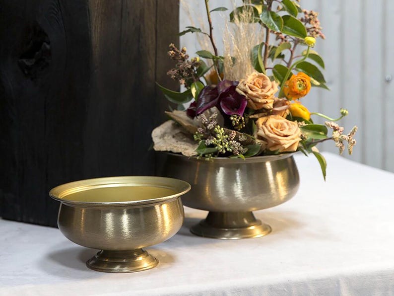 May include: Two antique brass bowls, one small and one large, sit on a white surface. The larger bowl holds a floral arrangement with peach roses, purple calla lilies, and greenery. The bowls have a textured finish and are on pedestal bases.