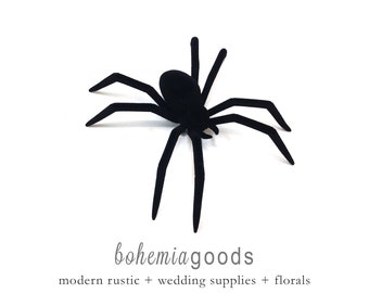 May include: A black spider decoration with eight legs and a rounded body, set against a white background. The spider is a Halloween or party decoration. The text "bohemiagoods" is below the spider, with the words "modern rustic + wedding supplies + florals" below that.