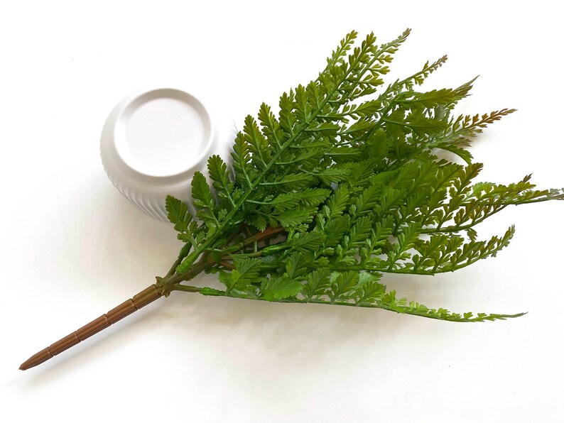 May include: A single stem of artificial green fern with a brown wire stem. The fern is arranged in a white ceramic pot.