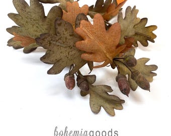 May include: A decorative branch featuring textured oak leaves in shades of muted green, brown, and orange, accented with several small brown acorns. The text "bohemiagoods" and "modern rustic + wedding supplies + florals" appears below the branch.