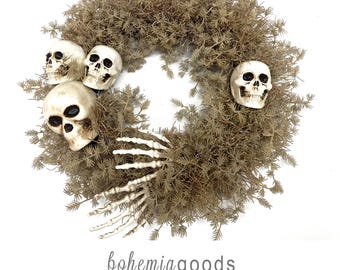 May include: A circular wreath made of light brown, needle-like foliage decorated with three off-white plastic human skulls and a white plastic skeletal hand. Below the wreath, the text reads: bohemiagoods modern rustic + wedding supplies + florals.