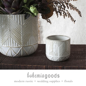 May include: Two white ceramic planters with a geometric design. The larger planter holds a floral arrangement with purple and green foliage. The smaller planter is empty. The text "bohemiagoods" is at the bottom, along with "modern rustic + wedding supplies + florals."
