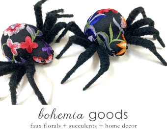 May include: Two plush spiders with black, furry bodies and embroidered floral designs on their heads. The spiders have multiple legs and are set against a white background. The text "bohemia goods" is at the bottom of the image.