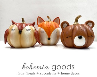 May include: Three ceramic pumpkin figurines with animal faces. The pumpkins are orange, brown, and cream colored. The animal faces are an owl, a fox, and a bear. Each pumpkin has a gold stem.