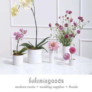 May include: White ceramic vases and pots display a variety of flowers, including orchids and dahlias. The flowers are in shades of pink, purple, and yellow. The text "bohemiagoods" and "modern rustic + wedding supplies + florals" are at the bottom.