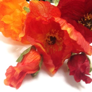 K&ouml;nnte beinhalten: Nahaufnahme von k&uuml;nstlichen Mohnblumen in Orange- und Rott&ouml;nen. Die Bl&uuml;ten haben detaillierte Bl&uuml;tenbl&auml;tter und dunkle Zentren, mit ein paar unge&ouml;ffneten Knospen. Das Arrangement ist vor einem wei&szlig;en Hintergrund platziert, der die leuchtenden Farben hervorhebt.