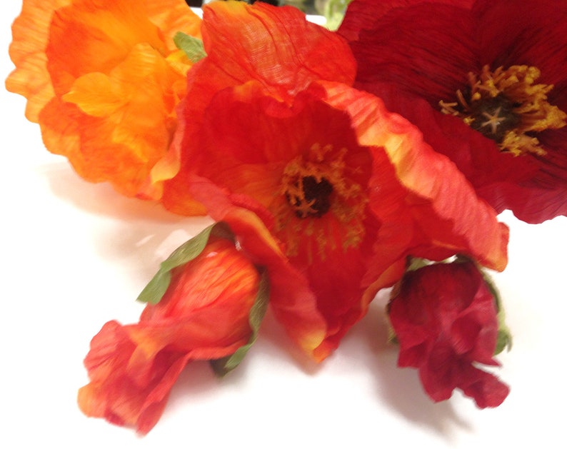 K&ouml;nnte beinhalten: Nahaufnahme von k&uuml;nstlichen Mohnblumen in Orange- und Rott&ouml;nen. Die Bl&uuml;ten haben detaillierte Bl&uuml;tenbl&auml;tter und dunkle Zentren, mit ein paar unge&ouml;ffneten Knospen. Das Arrangement ist vor einem wei&szlig;en Hintergrund platziert, der die leuchtenden Farben hervorhebt.
