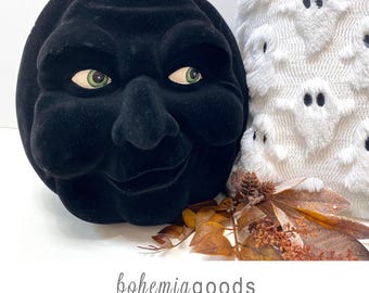May include: A black velvet pumpkin with a sculpted face featuring green eyes and a brown stem, positioned next to a white fabric item with a ghost pattern and brown dried leaves. The text bohemiagoods modern rustic + wedding supplies + florals.