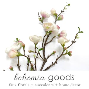 May include: A branch of white and pink faux flowers with green leaves. The branch is on a white background. The text "bohemia goods" is below the branch. The text "faux florals + succulents + home decor" is below the text "bohemia goods".