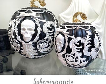 May include: Two black decorative pumpkins with white skull and scrollwork designs. The pumpkins have gold-colored stems and are part of a Halloween or gothic-themed collection. The pumpkins are on a glass surface.