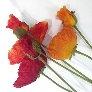 K&ouml;nnte beinhalten: Mehrere k&uuml;nstliche Mohnblumen in Rot- und Oranget&ouml;nen sind vor einem wei&szlig;en Hintergrund angeordnet. Die Bl&uuml;ten haben strukturierte Bl&uuml;tenbl&auml;tter und gr&uuml;ne Stiele. Die Mohnknospen befinden sich in verschiedenen Bl&uuml;hstadien.