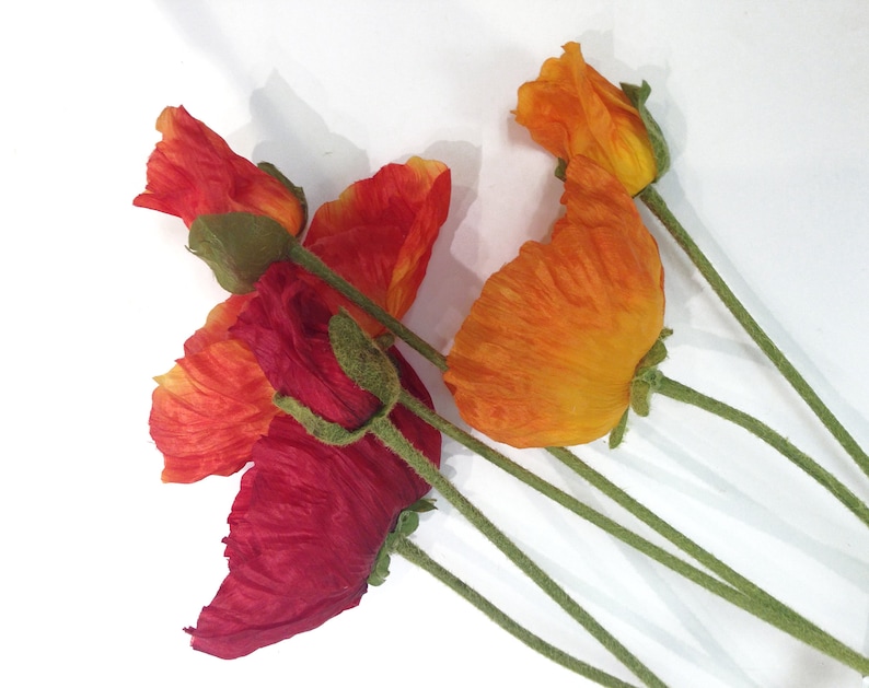 K&ouml;nnte beinhalten: Mehrere k&uuml;nstliche Mohnblumen in Rot- und Oranget&ouml;nen sind vor einem wei&szlig;en Hintergrund angeordnet. Die Bl&uuml;ten haben strukturierte Bl&uuml;tenbl&auml;tter und gr&uuml;ne Stiele. Die Mohnknospen befinden sich in verschiedenen Bl&uuml;hstadien.