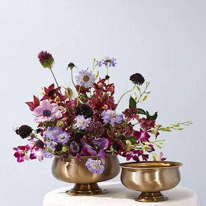 May include: A floral arrangement featuring a variety of flowers in shades of purple, burgundy, and white, displayed in two antique gold-colored bowls. The flowers include anemones, orchids, and scabiosa.