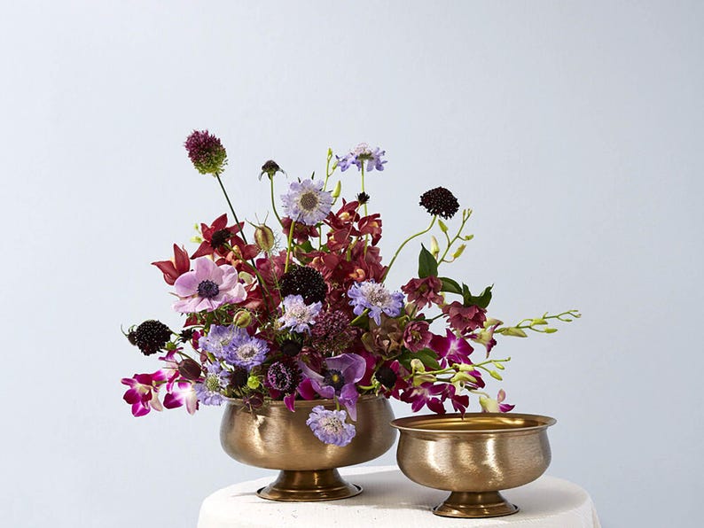 May include: A floral arrangement featuring a variety of flowers in shades of purple, burgundy, and white, displayed in two antique gold-colored bowls. The flowers include anemones, orchids, and scabiosa.