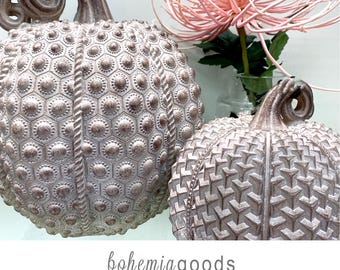 May include: Two decorative pumpkins with a metallic rose gold finish and textured surfaces, one featuring a hexagonal pattern with raised dots and the other a geometric chevron design, accompanied by a pink spider chrysanthemum flower.