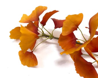 May include: A decorative branch featuring fan-shaped ginkgo leaves in vibrant shades of orange, yellow, and deep red. The leaves are attached to thin, flexible stems, showcasing a gradient of warm autumn colors with visible veining on each leaf.