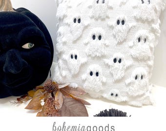May include: A square white throw pillow featuring a repeating pattern of raised, fuzzy ghost figures with black embroidered eyes. The text below reads bohemiagoods modern rustic + wedding supplies + florals in a simple gray sans-serif font.