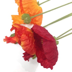 K&ouml;nnte beinhalten: Nahaufnahme von drei k&uuml;nstlichen Mohnblumen in Orange-, Rot- und Burgundert&ouml;nen. Die Bl&uuml;tenbl&auml;tter sind strukturiert und geschichtet, mit gr&uuml;nen Stielen und Knospen. Die Blumen sind vor einem wei&szlig;en Hintergrund angeordnet.