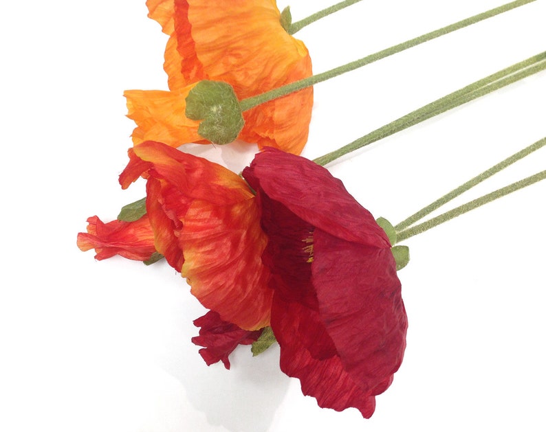 K&ouml;nnte beinhalten: Nahaufnahme von drei k&uuml;nstlichen Mohnblumen in Orange-, Rot- und Burgundert&ouml;nen. Die Bl&uuml;tenbl&auml;tter sind strukturiert und geschichtet, mit gr&uuml;nen Stielen und Knospen. Die Blumen sind vor einem wei&szlig;en Hintergrund angeordnet.