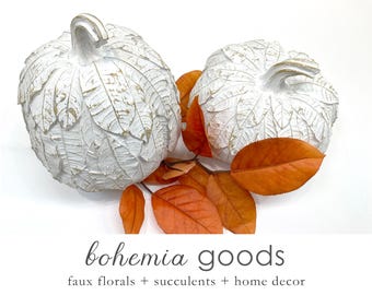May include: Two white decorative pumpkins with leaf-like textures and gold accents, resting among orange fall leaves. The pumpkins are part of a home decor collection, as indicated by the text "bohemia goods faux florals + succulents + home decor".