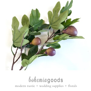 May include: Artificial fig branch with green leaves and ripe purple figs. The branch has brown stems and is set against a white background. The text "bohemiagoods" and "modern rustic + wedding supplies + florals" is at the bottom.